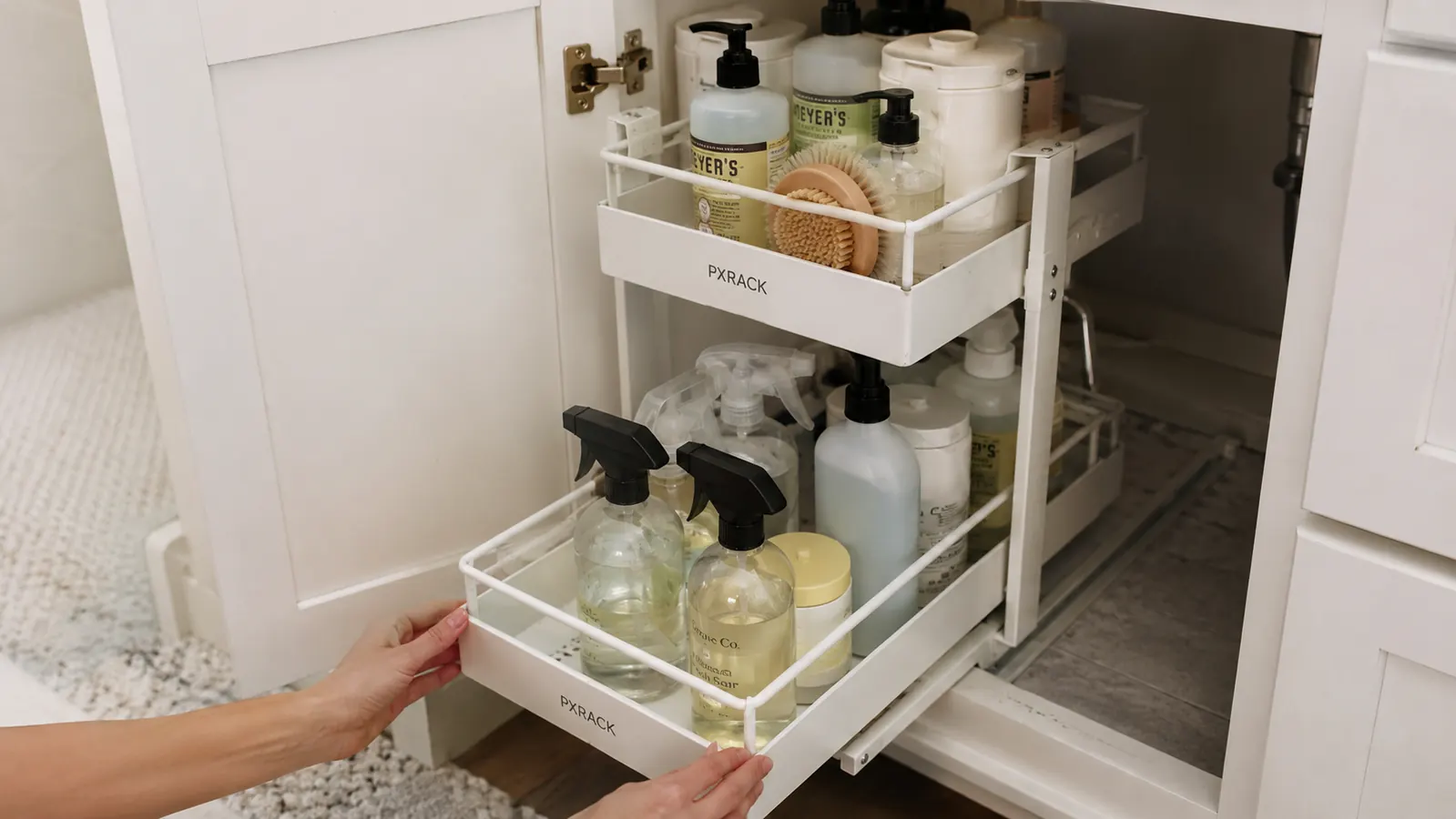Hands pulling out the lower tier of a white PXRACK under-sink organizer loaded with spray bottles and cleaning supplies